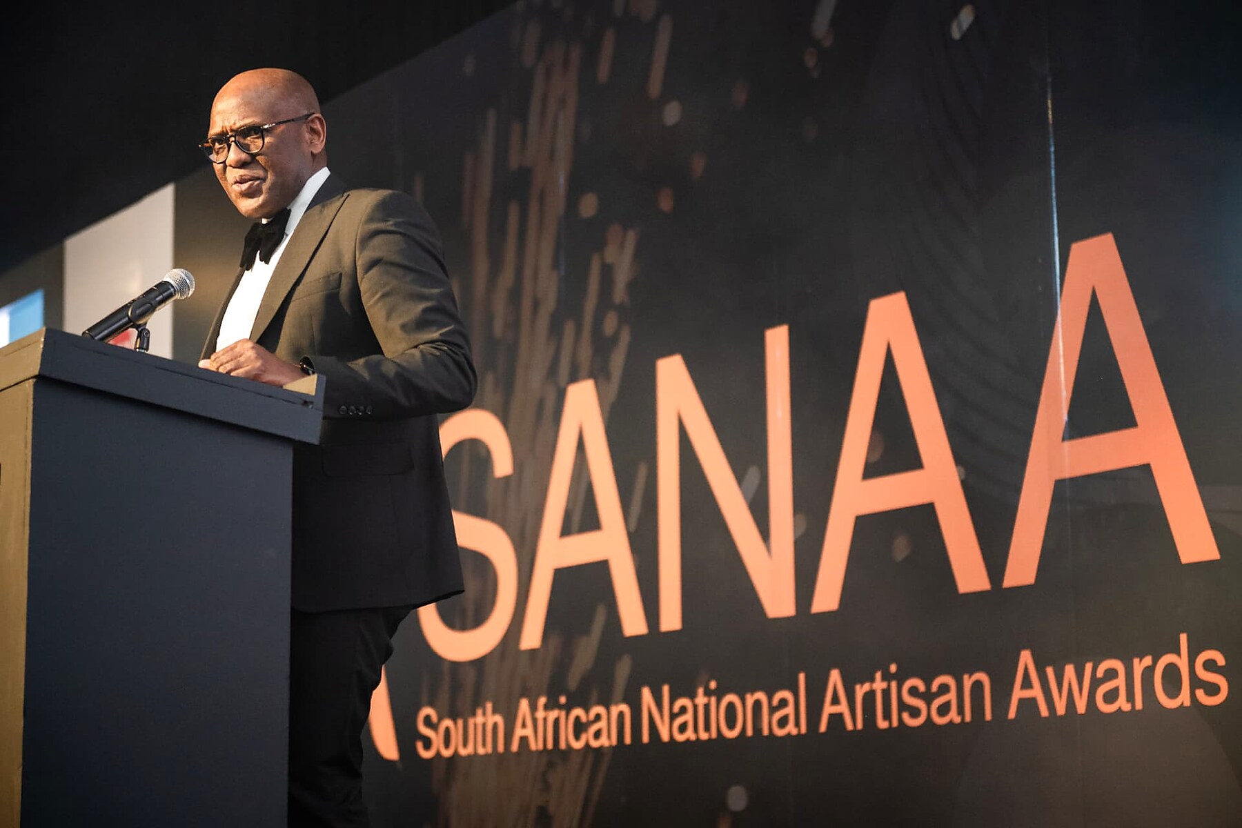 A speaker addressing the audience from the podium in front of the SANAA backdrop