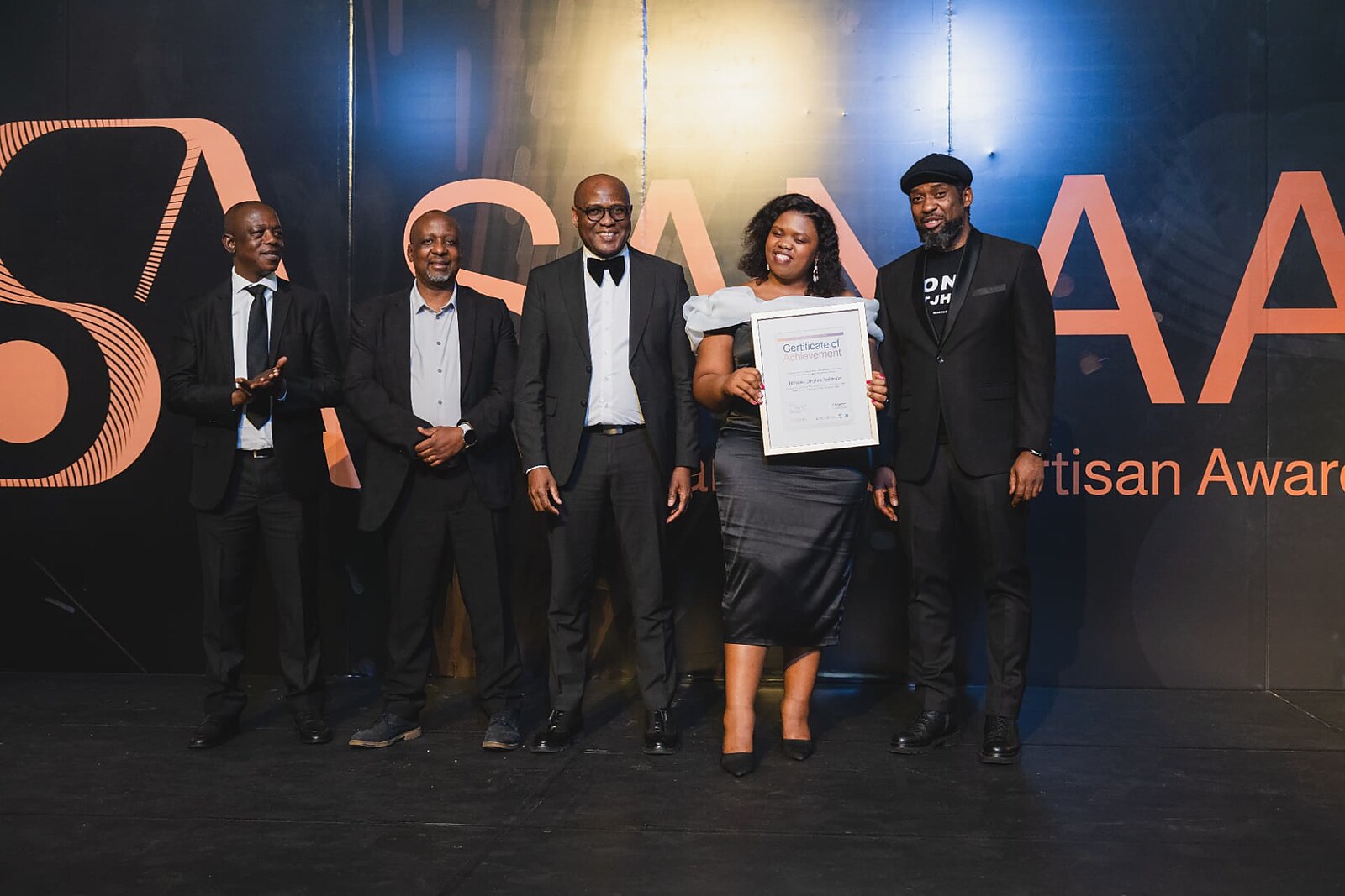 Female winner in an off-shoulder gown holding her framed Certificate of Achievement as officials applaud