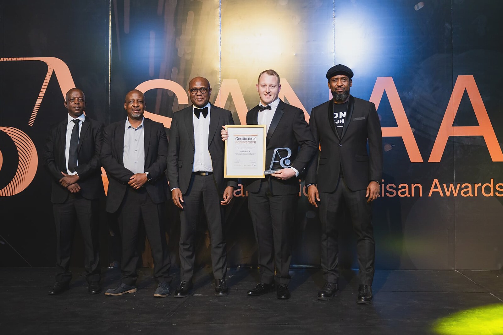 Winner in a bow tie holding his Certificate of Achievement and glass trophy with four officials on the SANAA stage