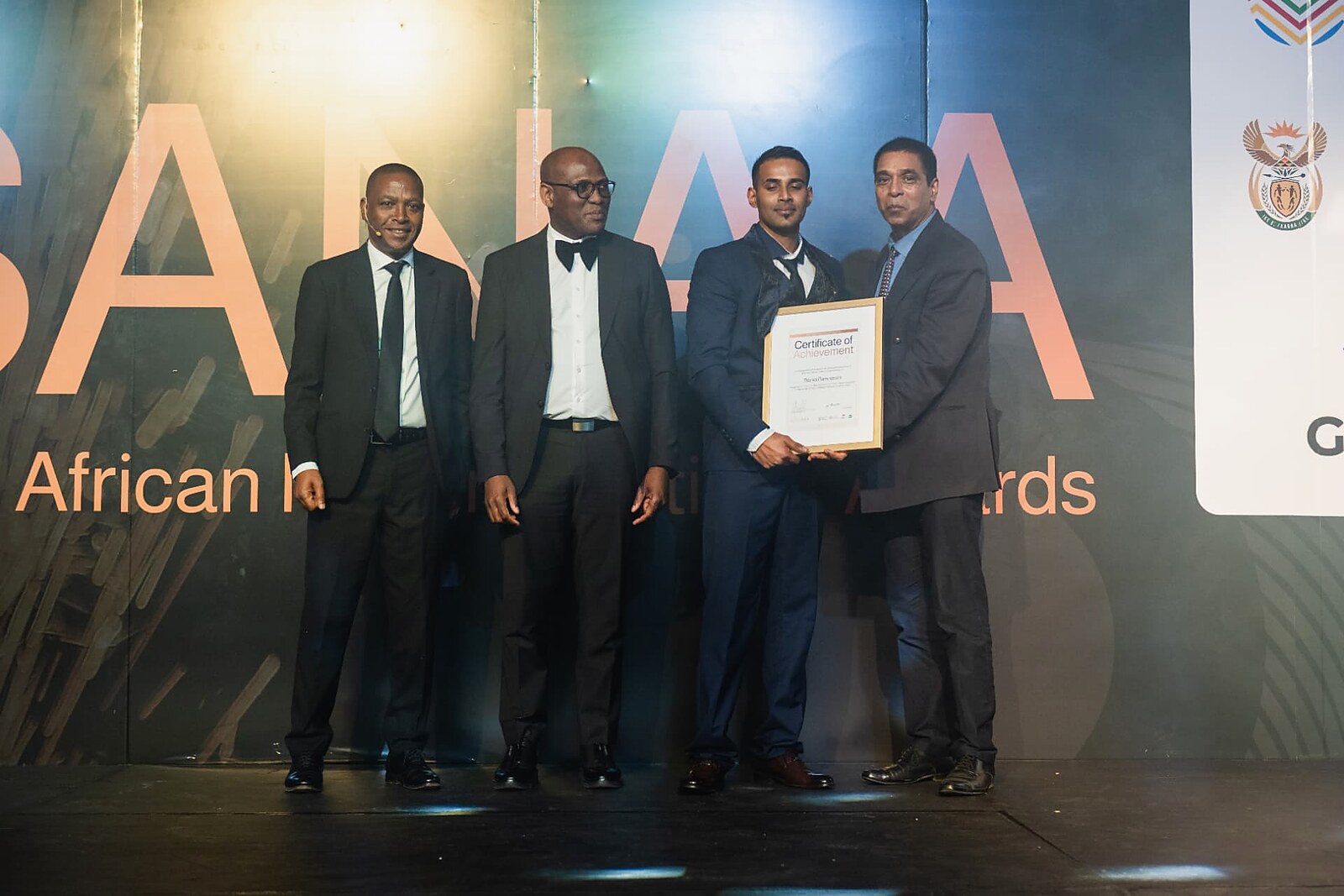Winner in a navy suit receiving his framed Certificate of Achievement from the awards panel