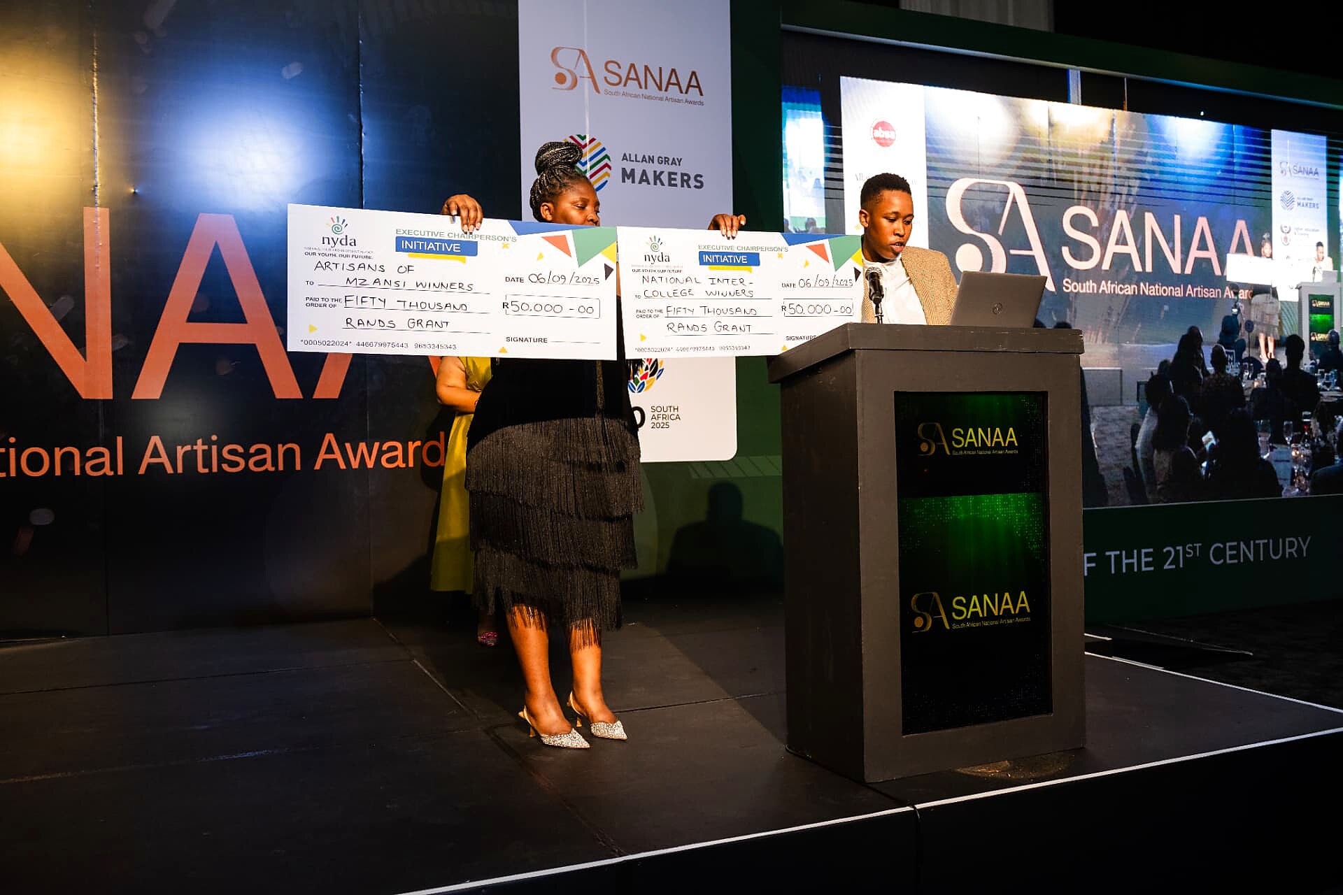 Two officials on stage holding oversized NYDA R50,000 grant cheques for the Artisans of Mzansi and National Inter-College winners