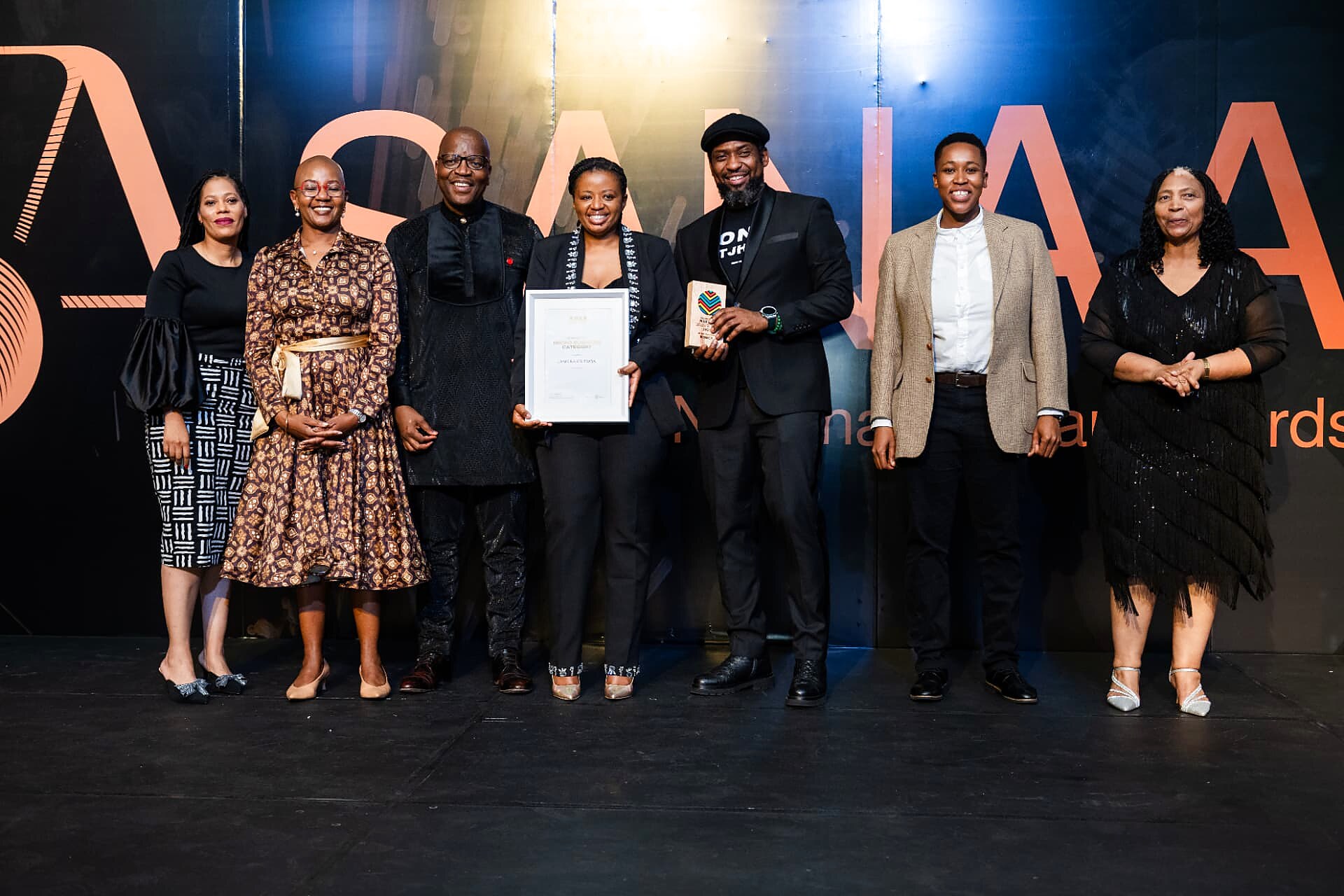 A winner holding her framed certificate and trophy on stage with the SANAA panel
