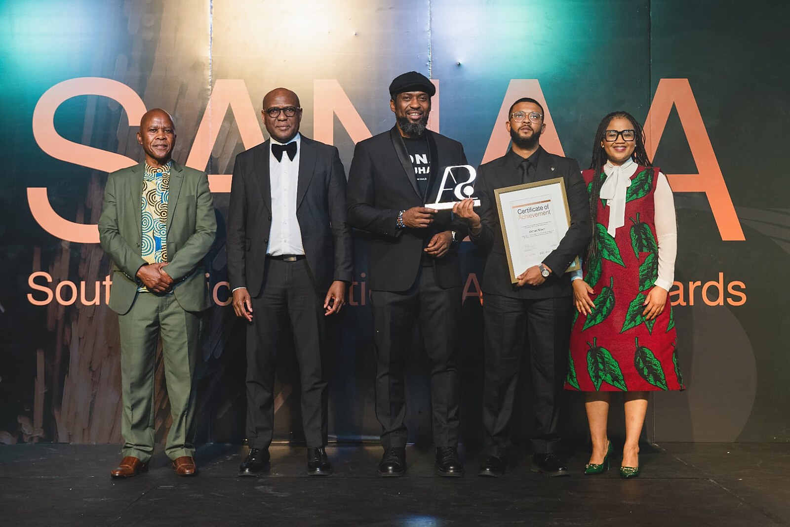 Winner in a black suit holding his framed Certificate of Achievement beside the host with the SA trophy on the SANAA stage