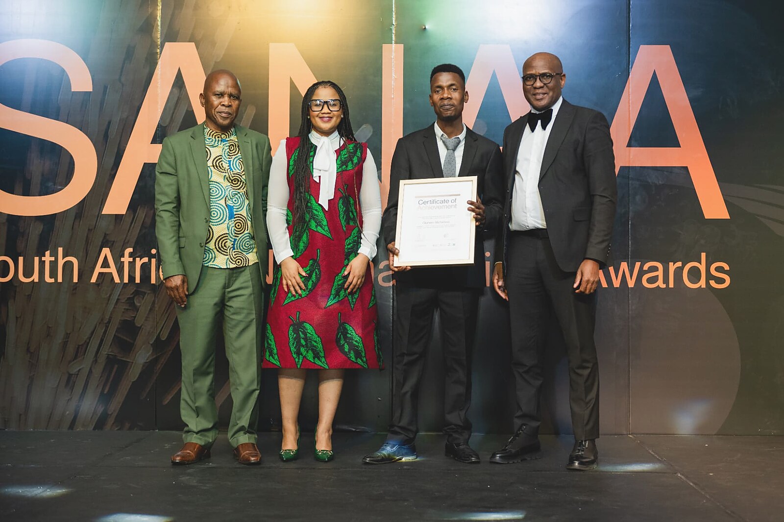 Winner in a dark suit holding his framed Certificate of Achievement with three SANAA officials on stage