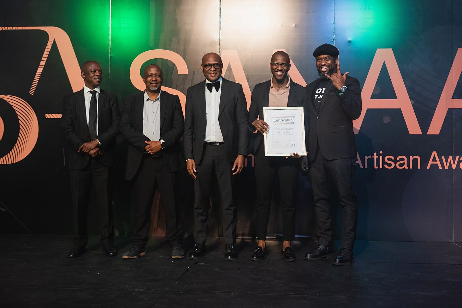 Recipient holding his Certificate of Achievement on stage under green and blue gala lighting