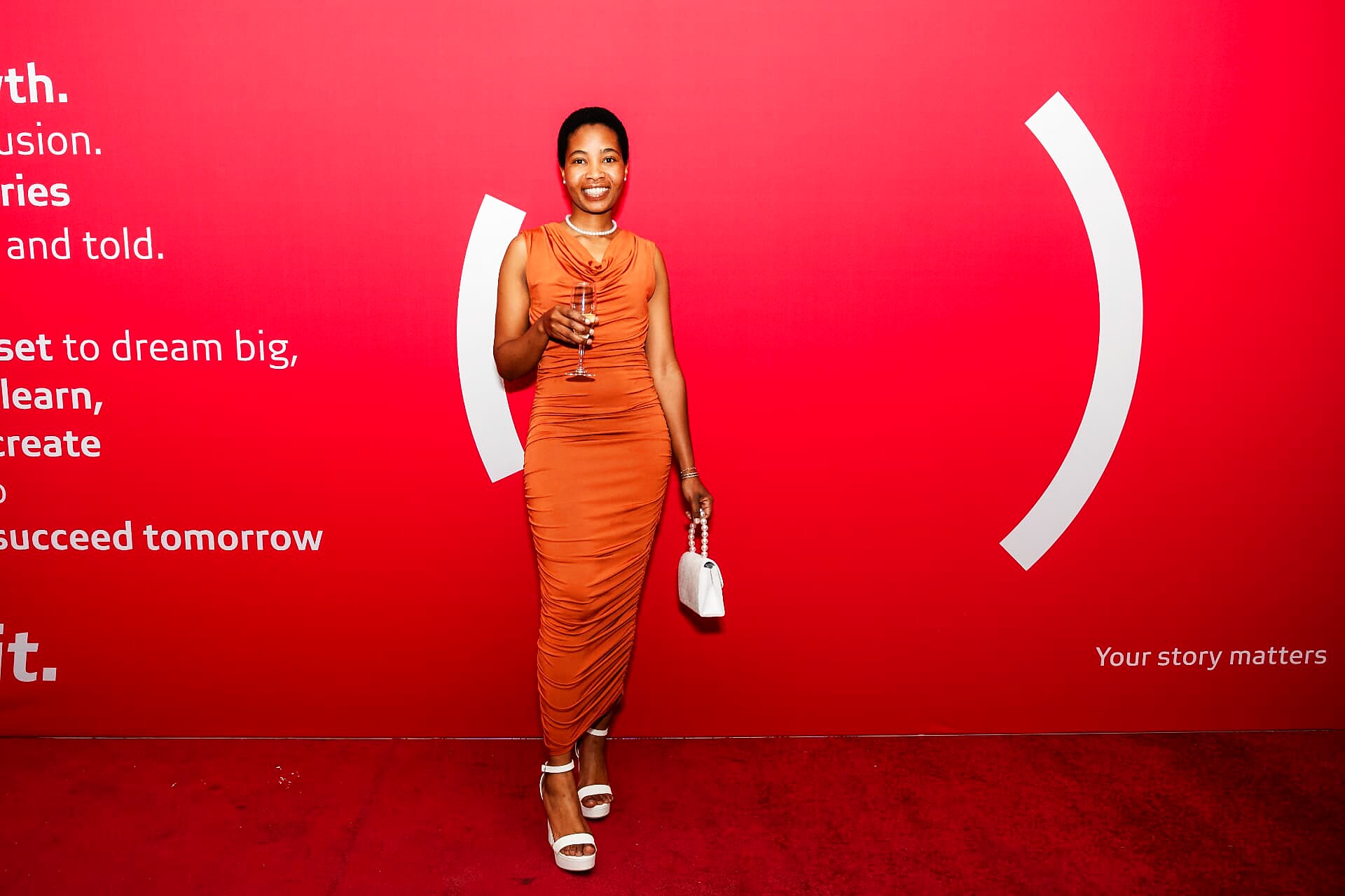 Guest in an orange gown on the SANAA 2026 red carpet holding a glass of sparkling wine