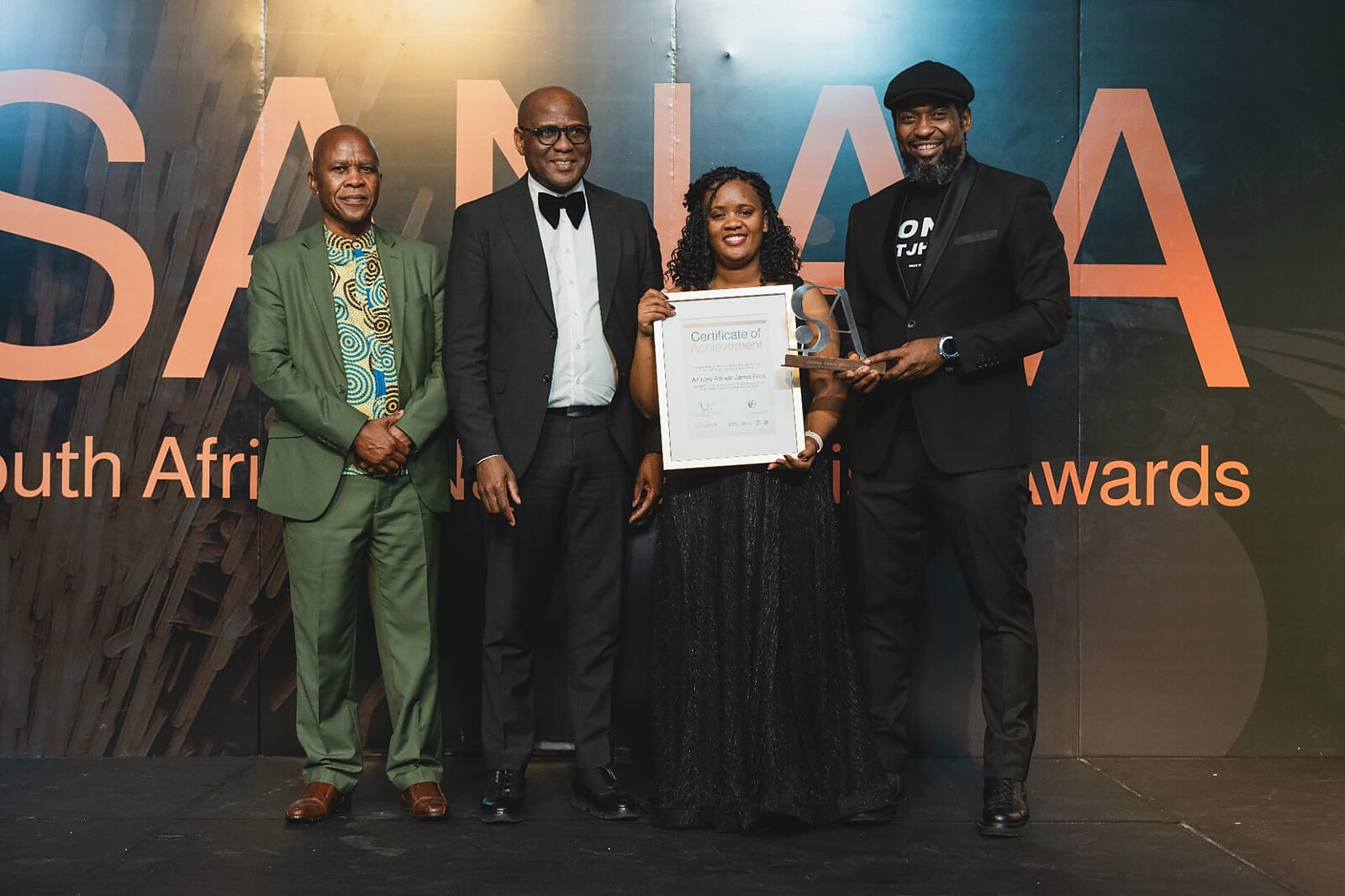 Winner in a black gown holding her framed Certificate of Achievement and trophy with officials on stage