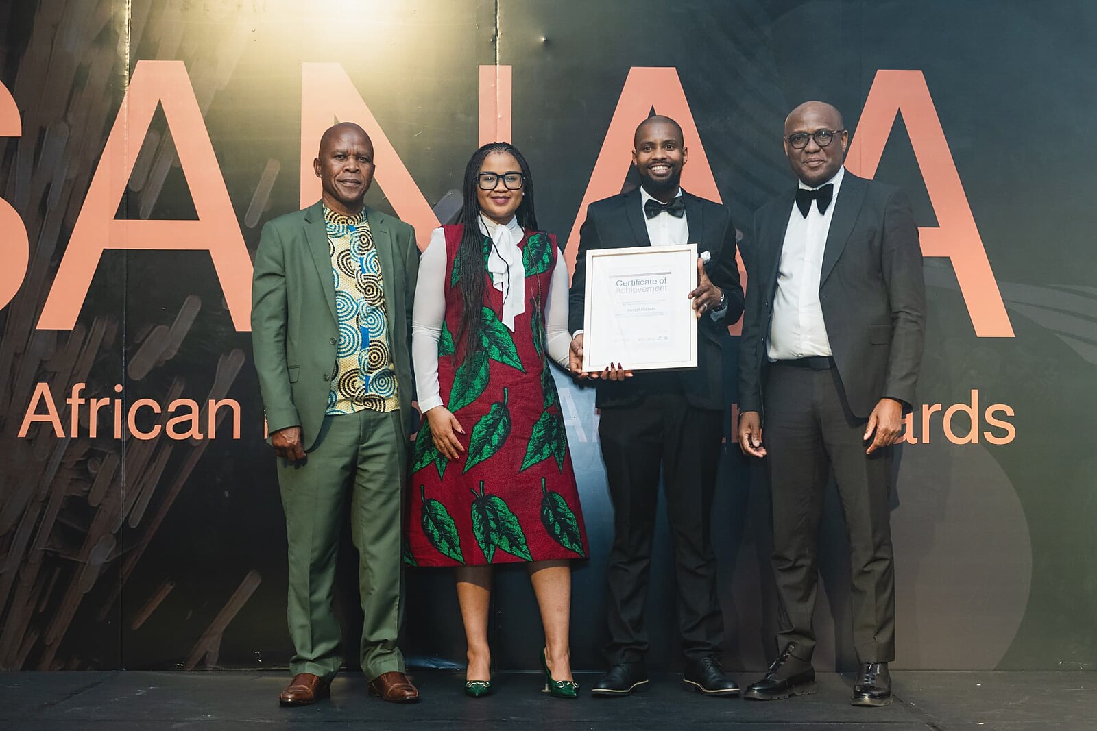 Winner in a bow tie and tuxedo holding his framed certificate with SANAA officials