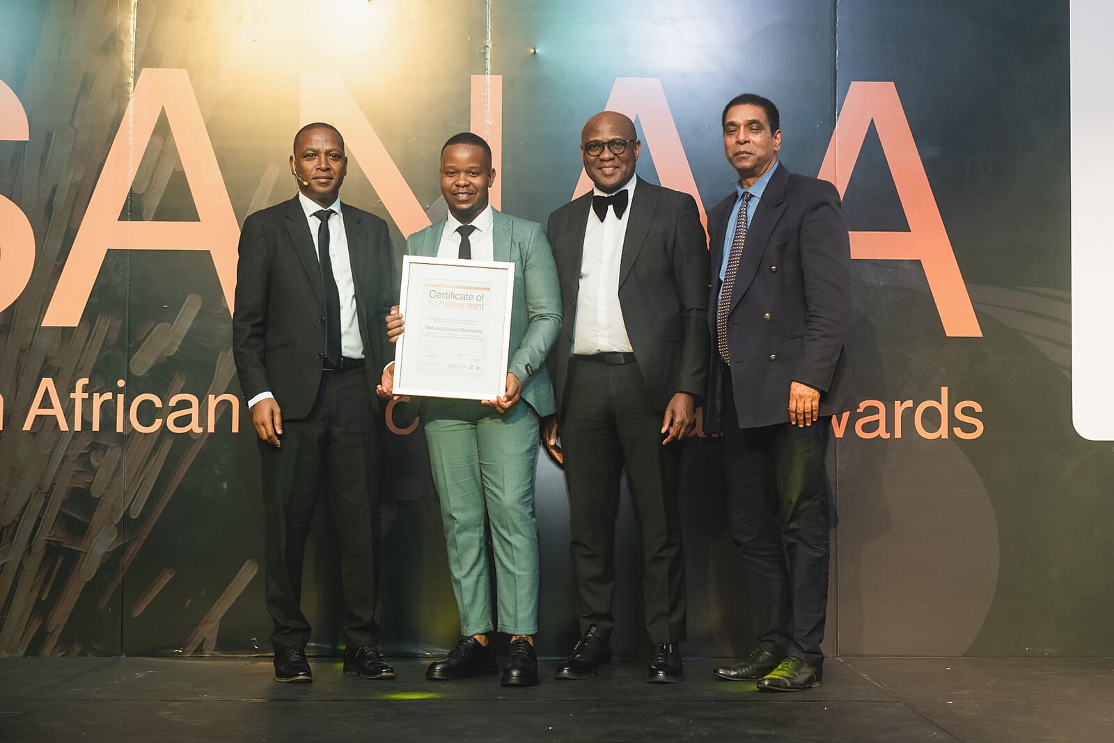 Winner in a sage green suit holding his framed Certificate of Achievement with the awards panel