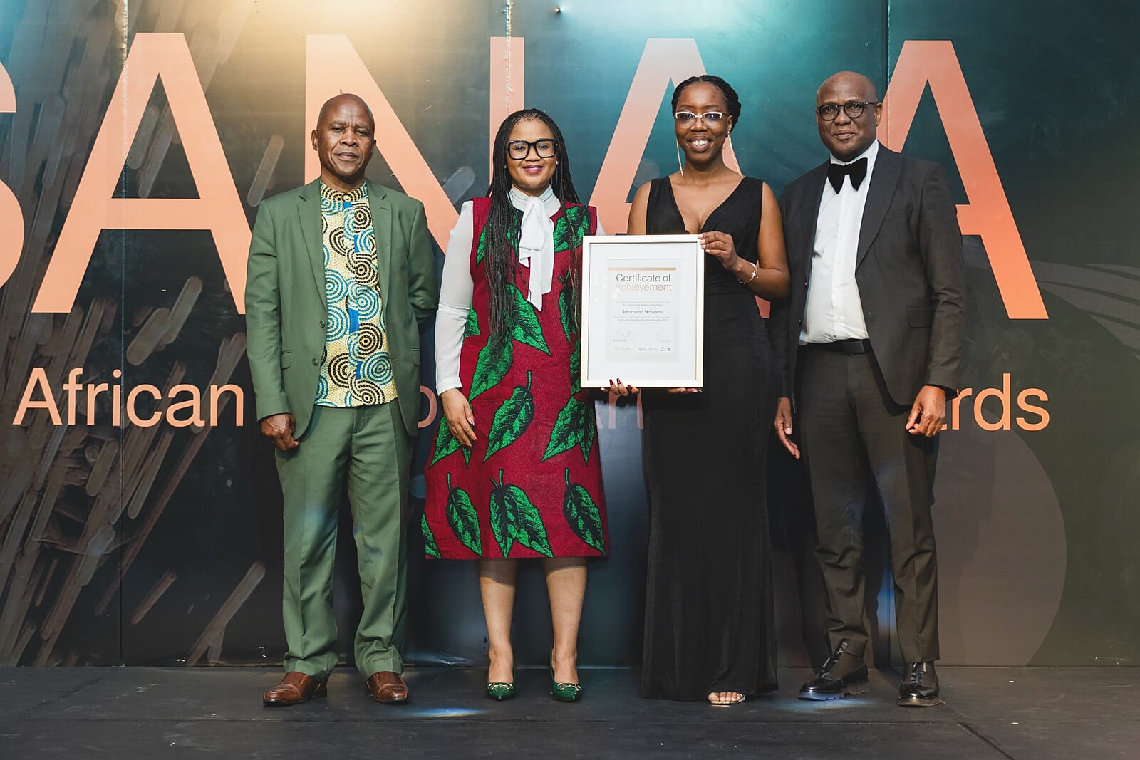 Female winner in a black evening gown holding her framed Certificate of Achievement on stage