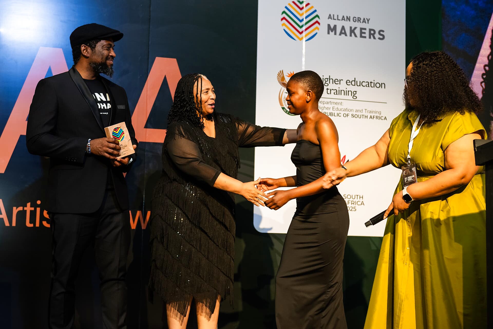 A winner being congratulated with a handshake on stage as her Allan Gray Makers trophy is presented