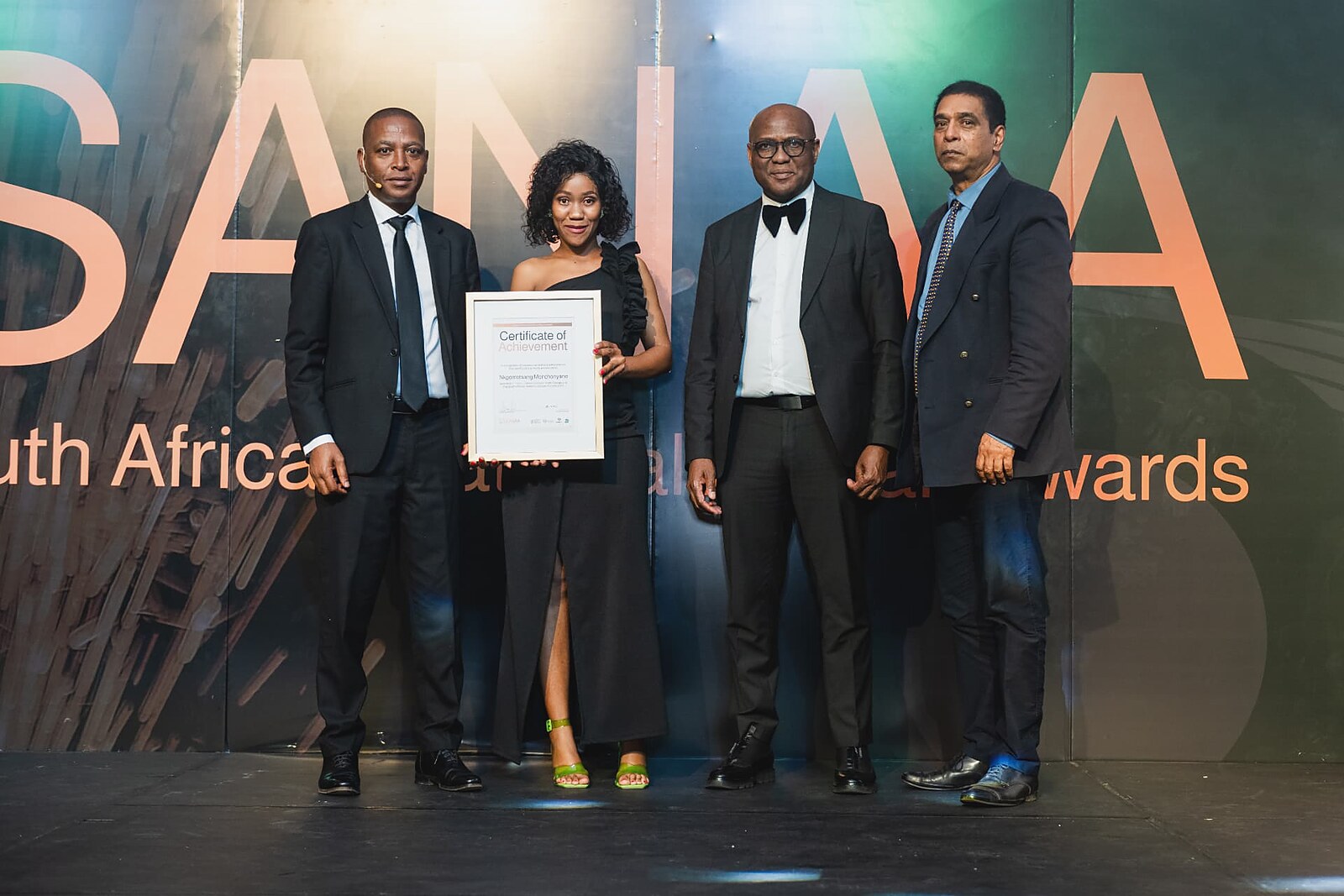 Female recipient in a black gown holding her framed certificate with the host and dignitaries