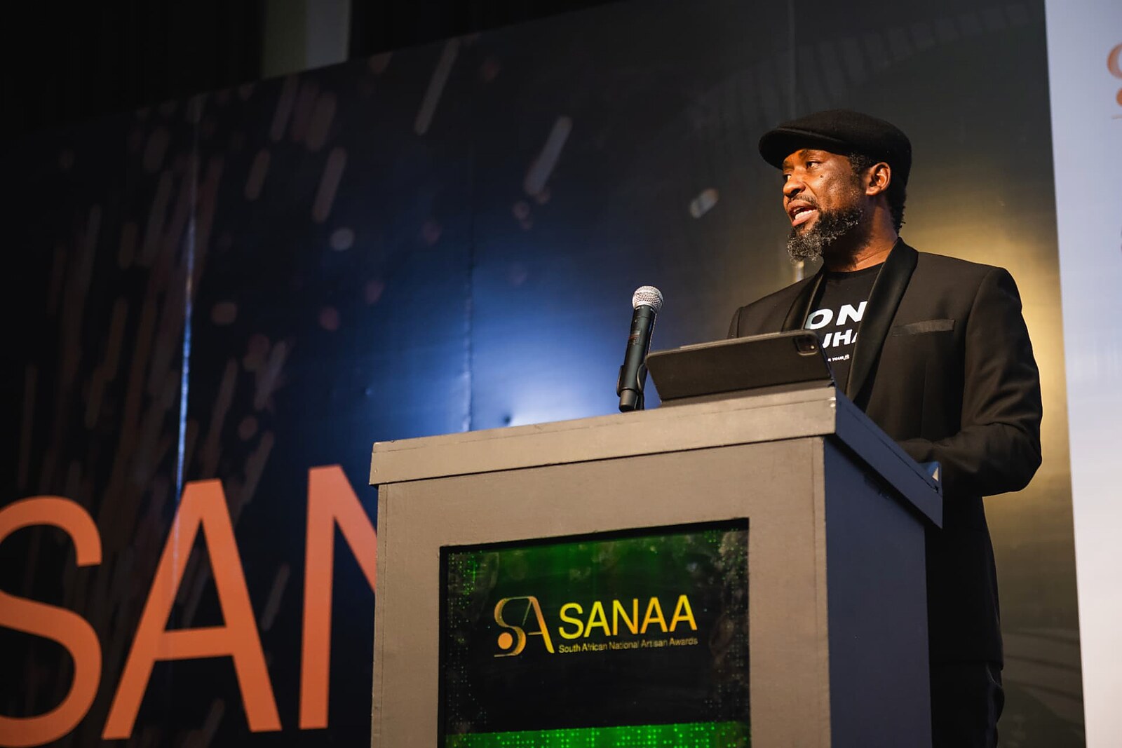 Host speaking at the podium with the SANAA branding behind him
