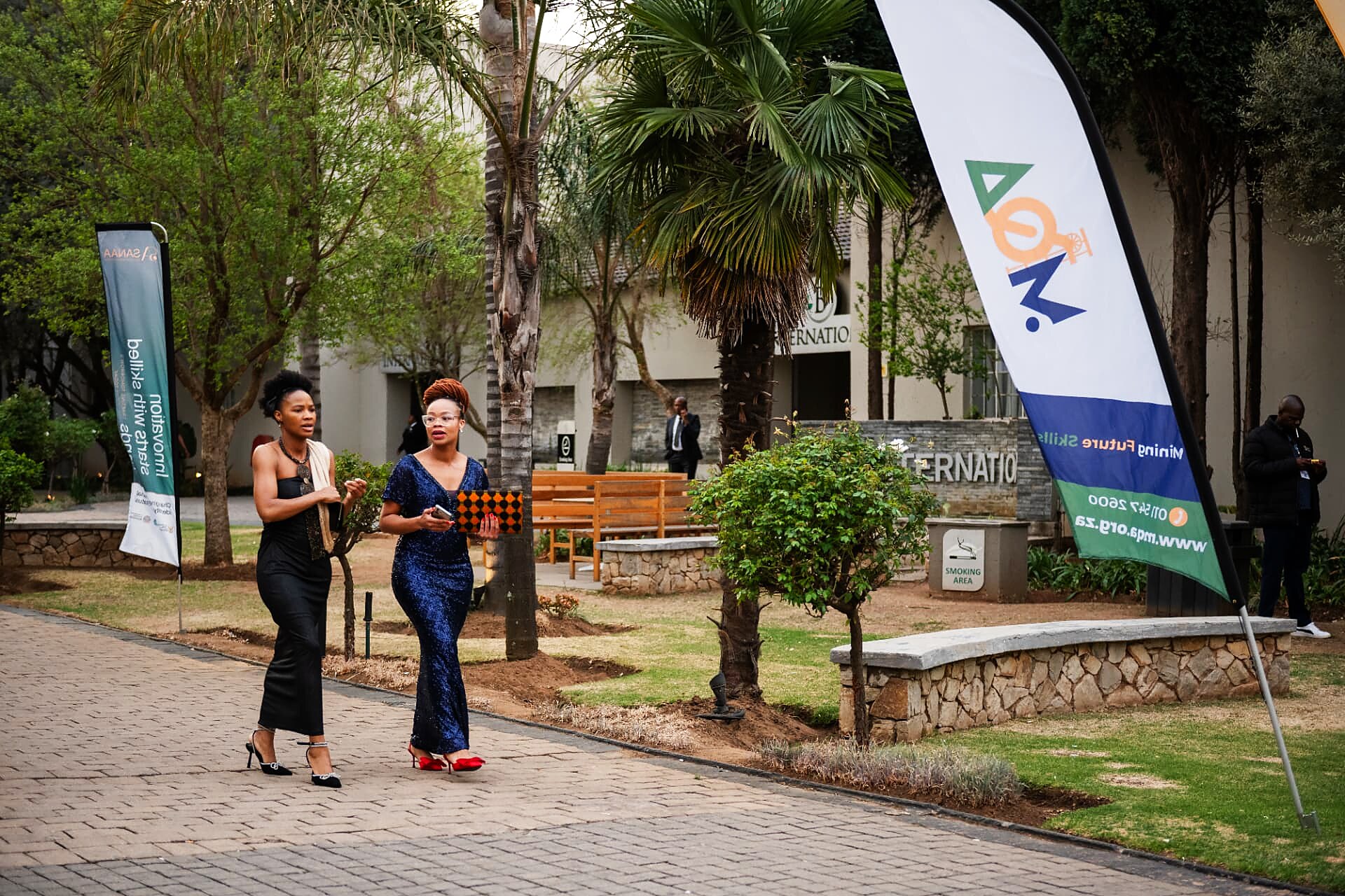 Two guests in evening gowns walking past SANAA and sponsor flags at the venue entrance