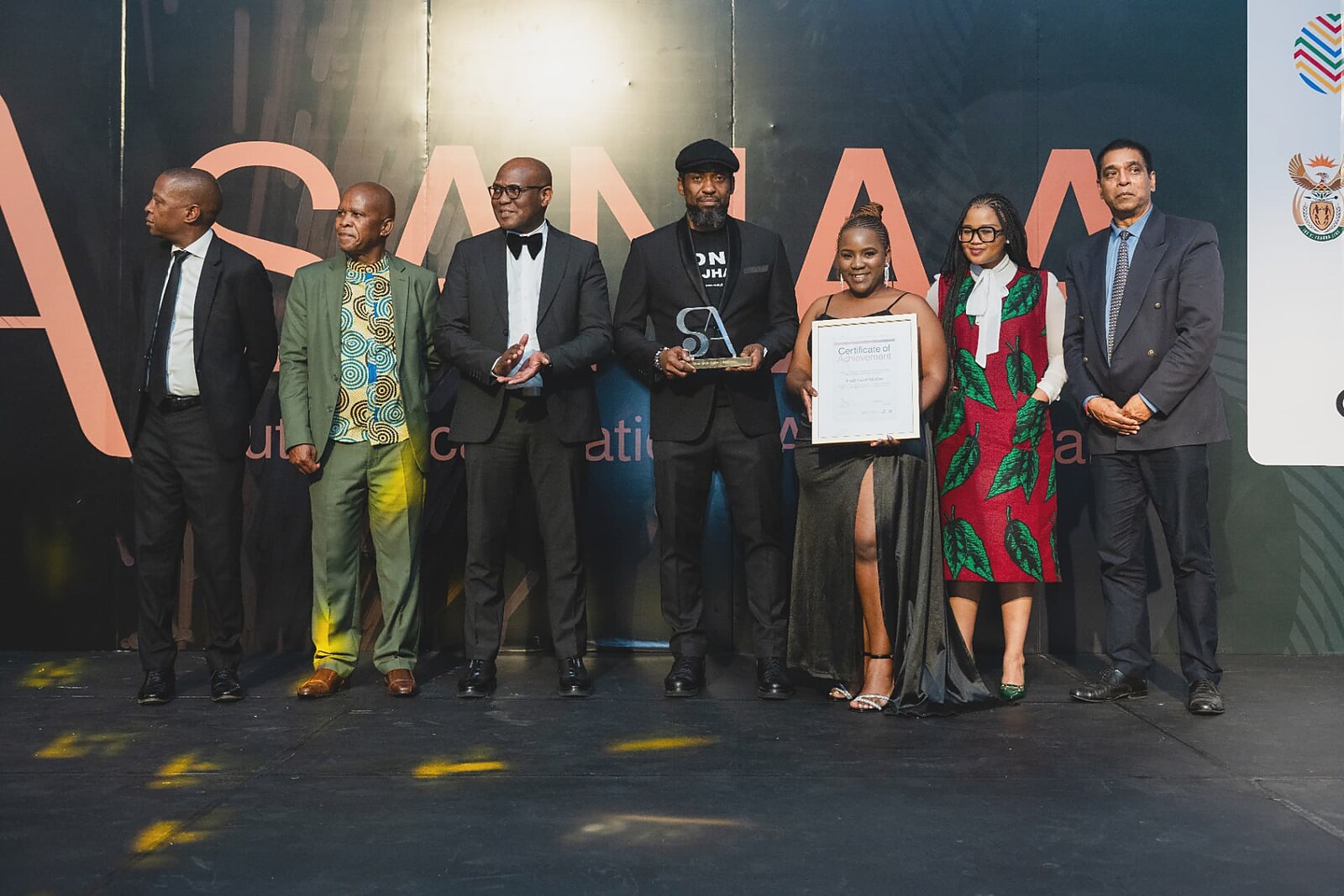 Female winner in a black gown holding her framed certificate while the host presents the glass SA trophy