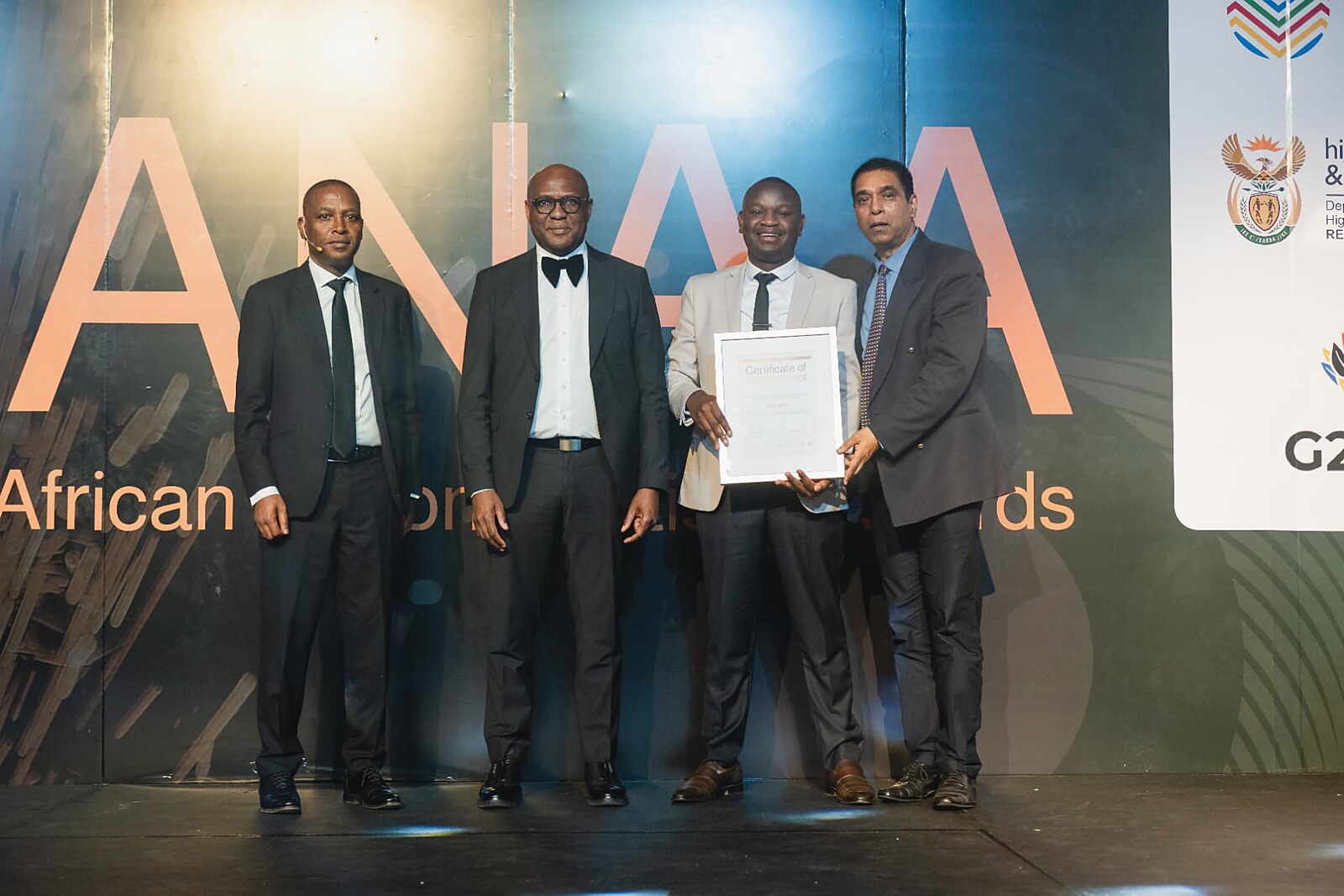 Winner in a light grey suit receiving his framed Certificate of Achievement from officials on the SANAA stage
