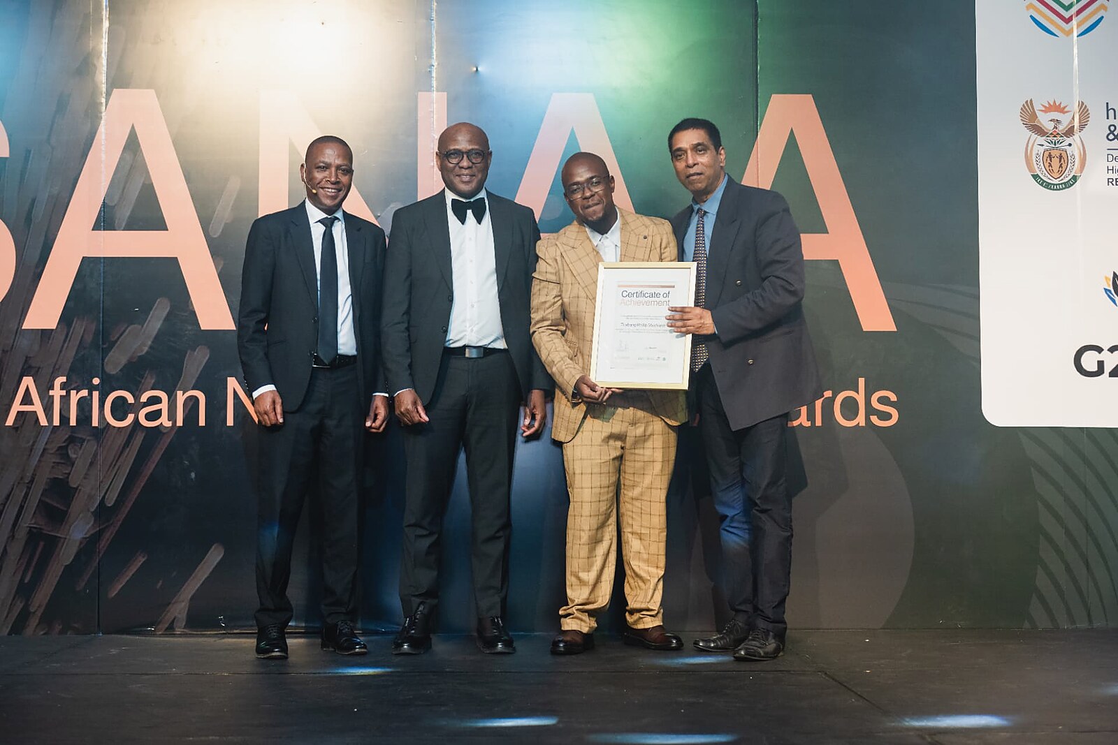 Winner in a tan check suit holding his framed Certificate of Achievement with the SANAA panel