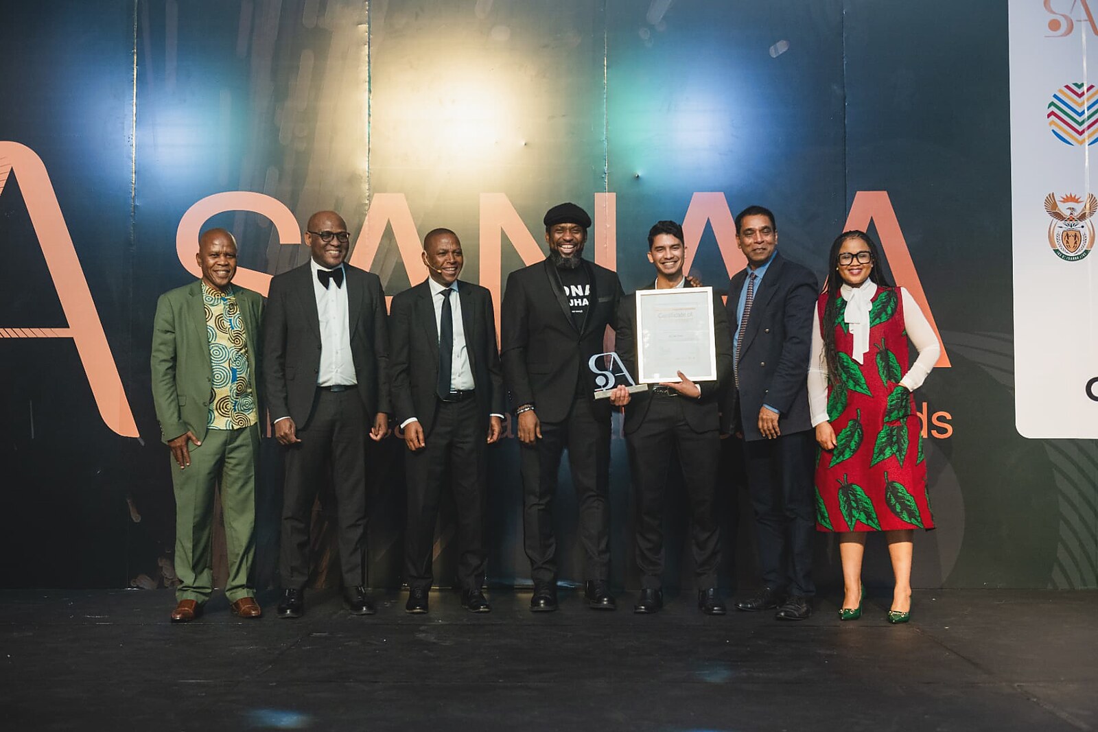 Winner holding his framed certificate and gold SA trophy with departmental officials in front of the SANAA wall
