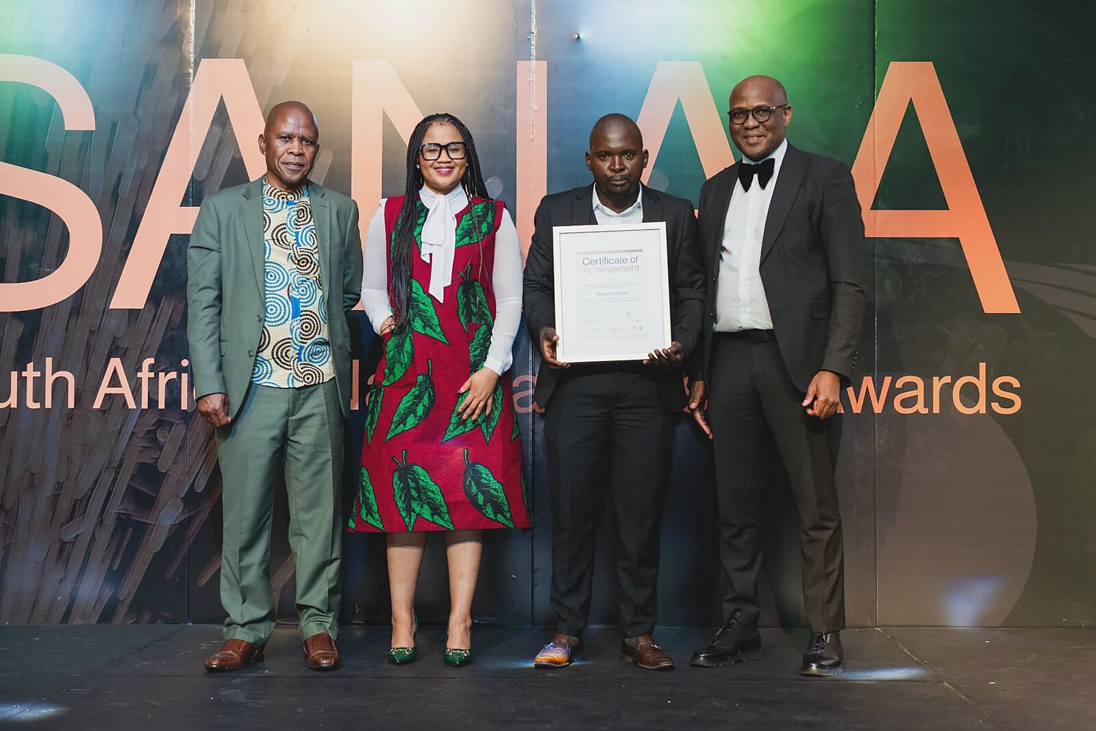 Award recipient presenting his framed Certificate of Achievement flanked by the SANAA leadership team
