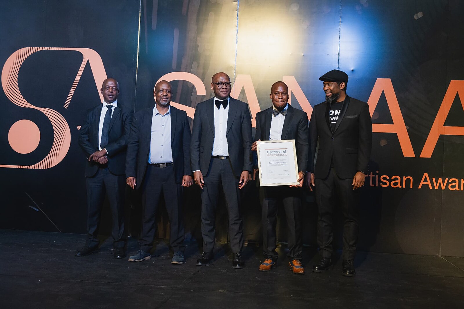 Award recipient holding his framed certificate on stage while the SANAA leadership team smiles beside him