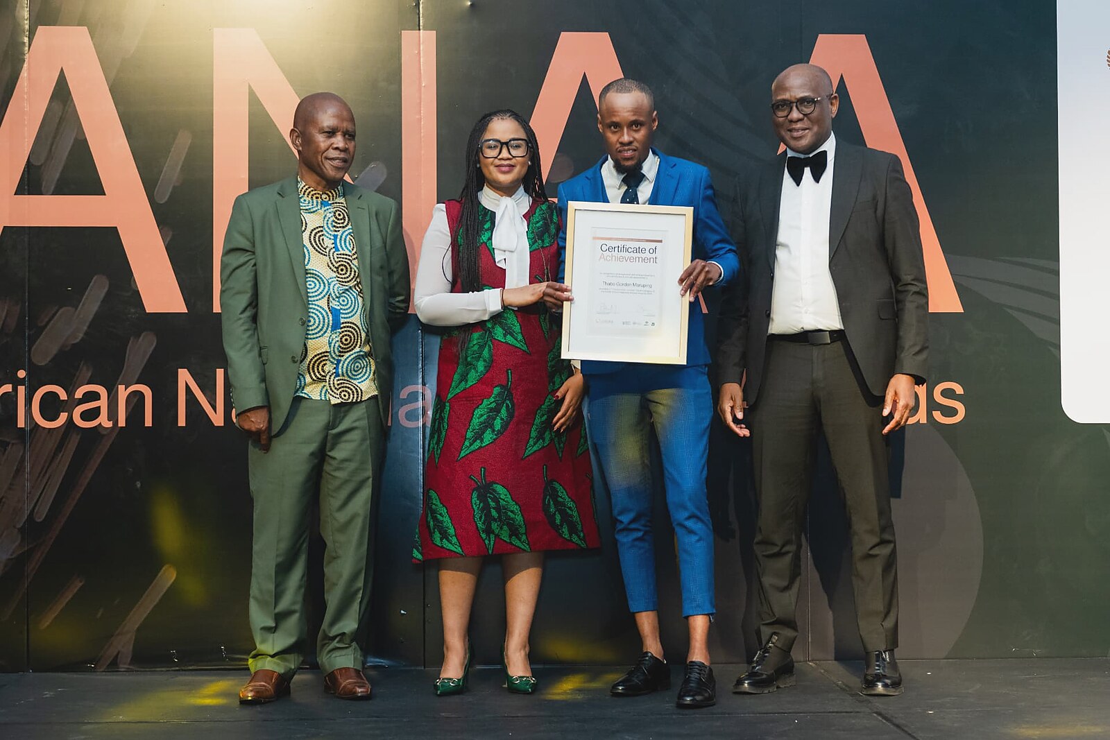 Winner in a blue check suit receiving his framed Certificate of Achievement on the SANAA stage