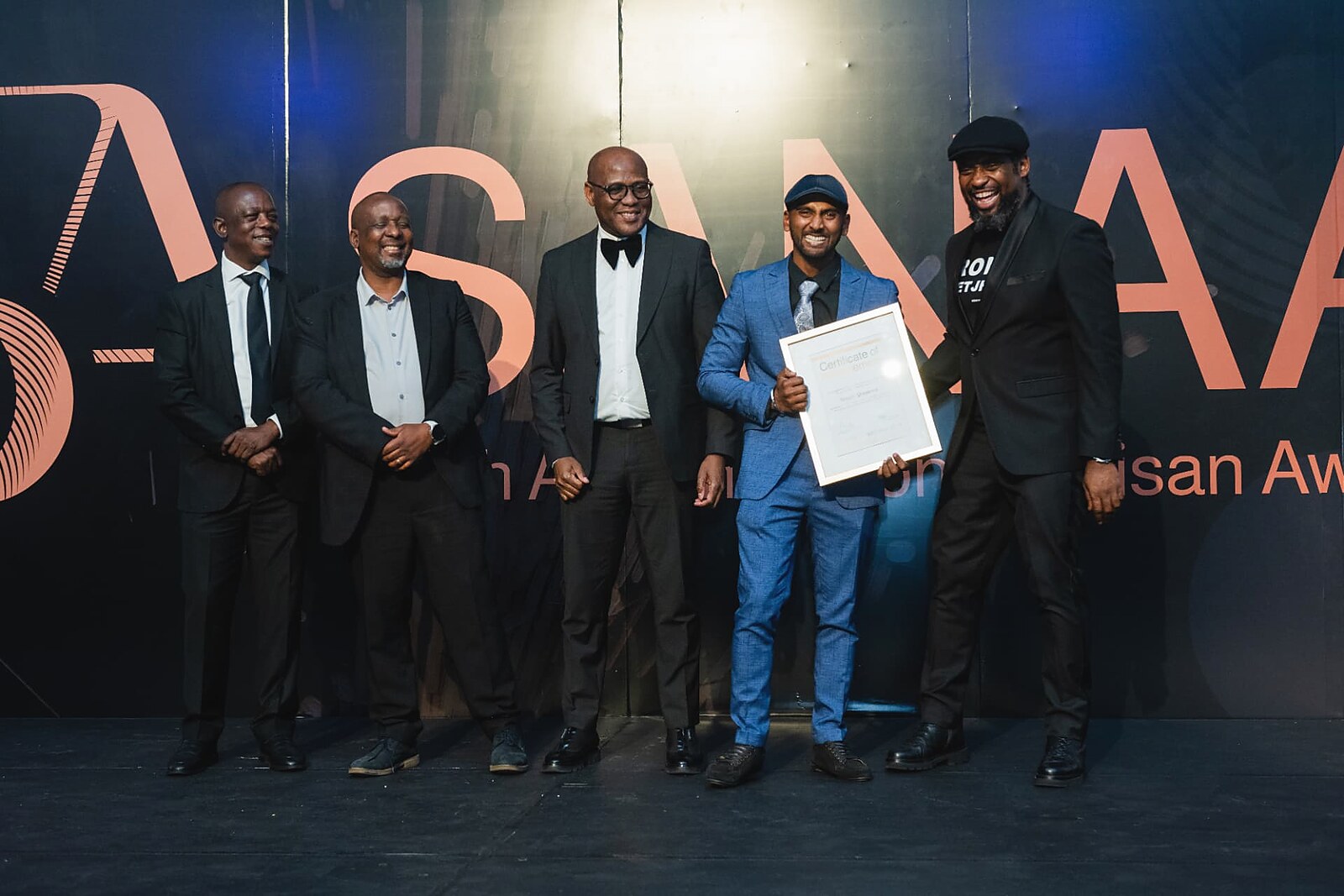 Winner in a blue suit laughing with the SANAA panel while holding his framed certificate
