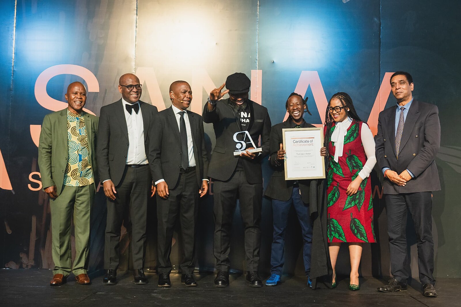 Winner celebrating on stage with his framed certificate as the host raises a hand in salute
