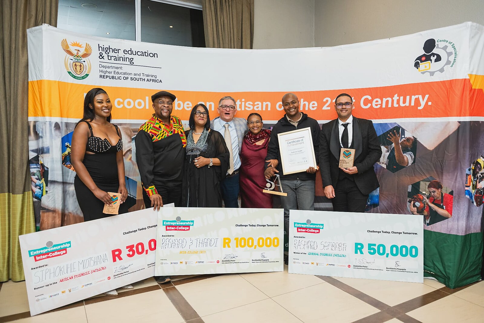 Group of winners holding Entrepreneurship Inter-College cheques in front of the Artisan of the 21st Century banner