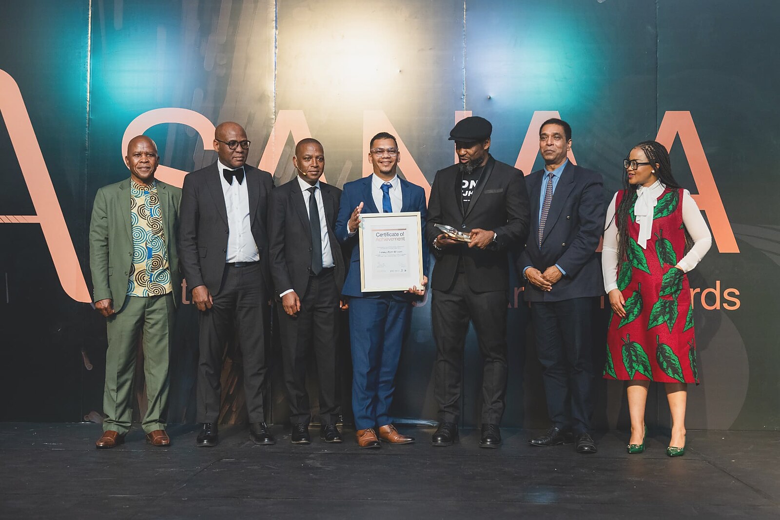 Winner in a blue suit presenting his framed Certificate of Achievement alongside the awards panel