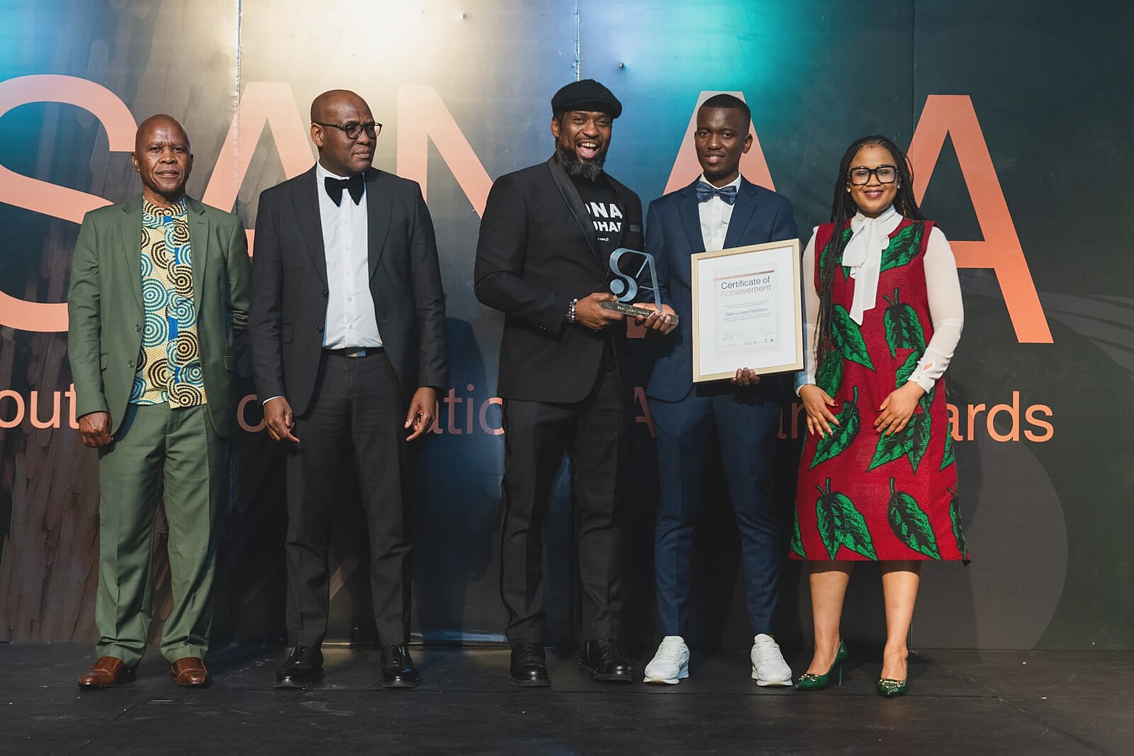Young artisan in a navy suit and bow tie receiving his Certificate of Achievement with the SANAA panel