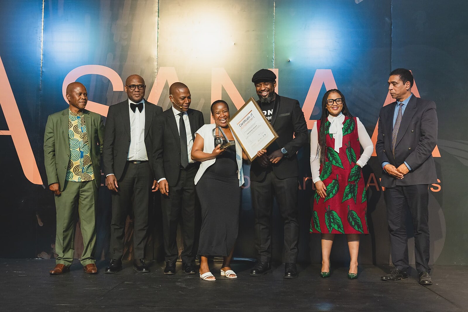 Laughing winner holding her framed certificate and SA trophy surrounded by the SANAA leadership team